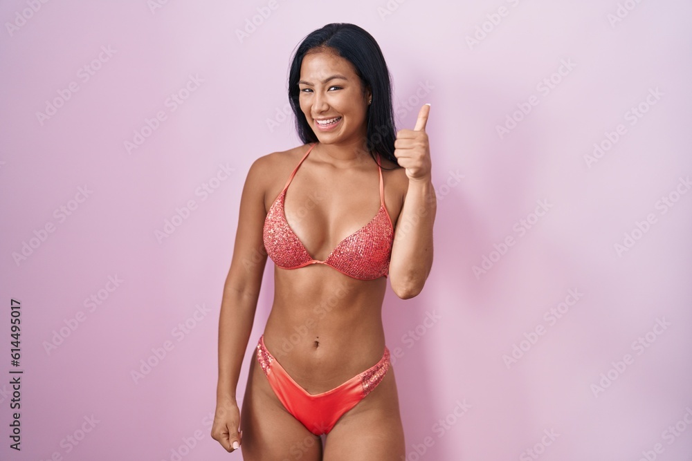 Hispanic woman wearing bikini doing happy thumbs up gesture with hand. approving expression looking at the camera showing success.