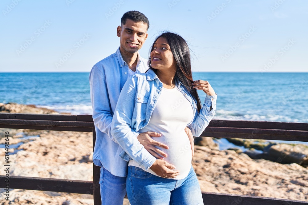 Young latin couple expecting baby hugging each other standing at seaside