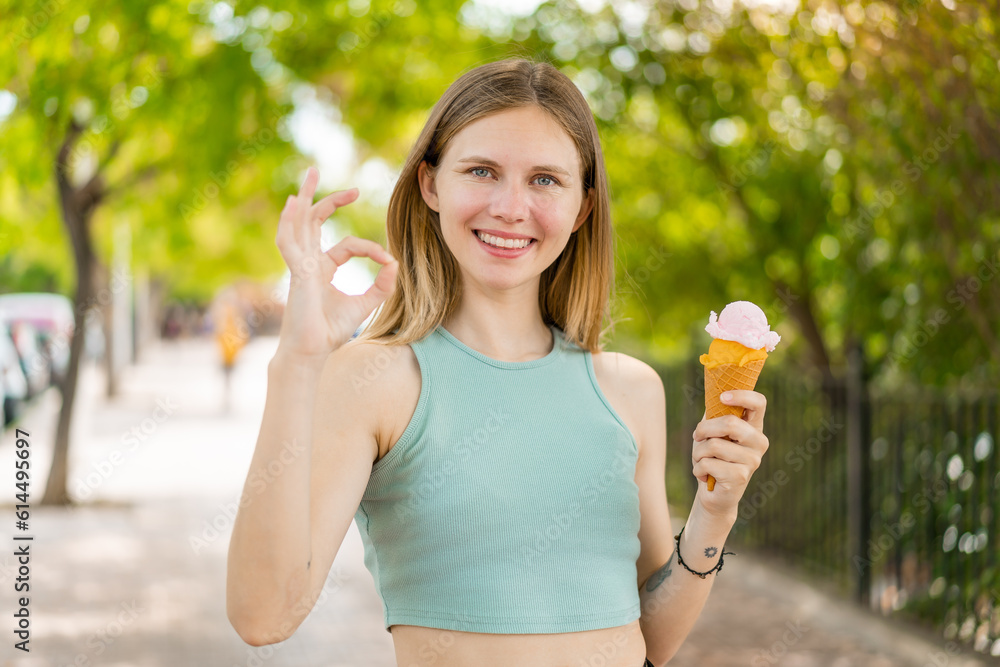 Young blonde pretty woman with a cornet ice cream at outdoors showing ok sign with fingers