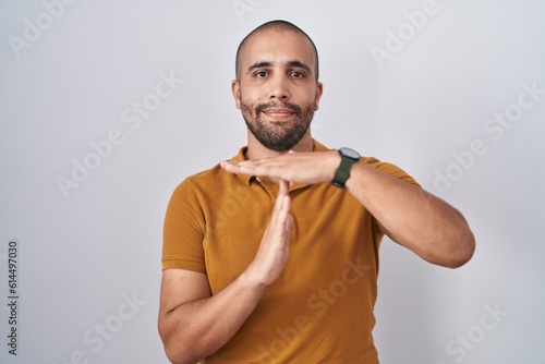 Hispanic man with beard standing over white background doing time out gesture with hands, frustrated and serious face