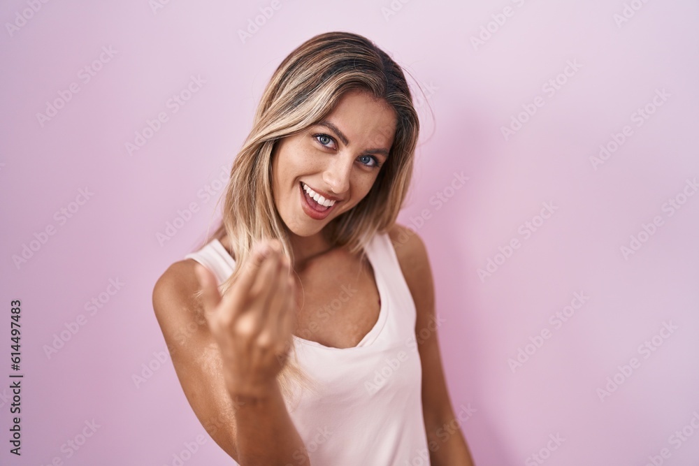 Young blonde woman standing over pink background beckoning come here gesture with hand inviting welcoming happy and smiling Young blonde woman standing over pink background beckoning come here gesture with hand inviting welcoming happy and smiling