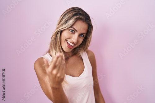 Young blonde woman standing over pink background beckoning come here gesture with hand inviting welcoming happy and smiling