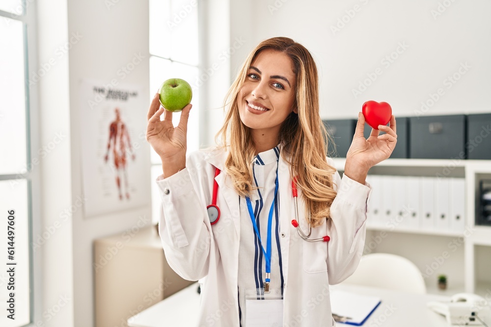 Young blonde doctor woman holding heart and green apple smiling with a happy and cool smile on face. showing teeth. Young blonde doctor woman holding heart and green apple smiling with a happy and cool smile on face. showing teeth.