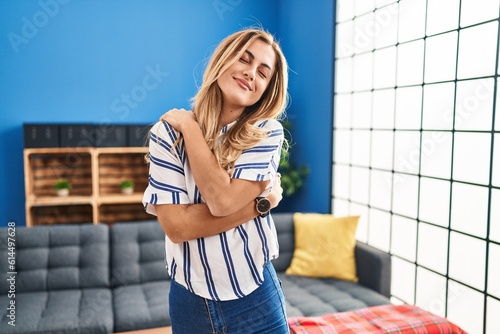 Canvastavla Young blonde woman standing at living room at home hugging oneself happy and positive, smiling confident