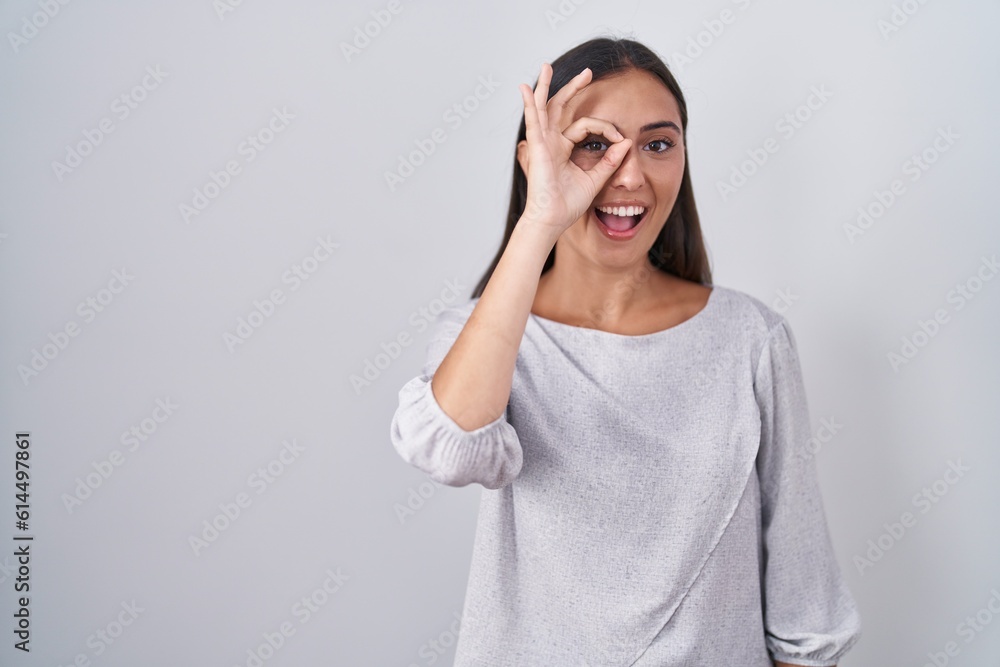 Young hispanic woman standing over white background doing ok gesture with hand smiling, eye looking through fingers with happy face. Young hispanic woman standing over white background doing ok gesture with hand smiling, eye looking through fingers with happy face.
