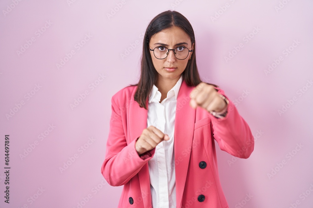 Young hispanic woman wearing business clothes and glasses punching fist to fight, aggressive and angry attack, threat and violence