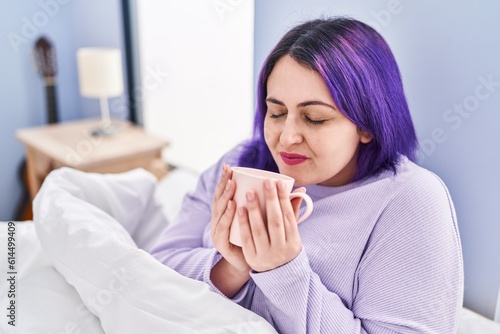 Young beautiful plus size woman drinking cup of coffee sitting on bed at bedroom