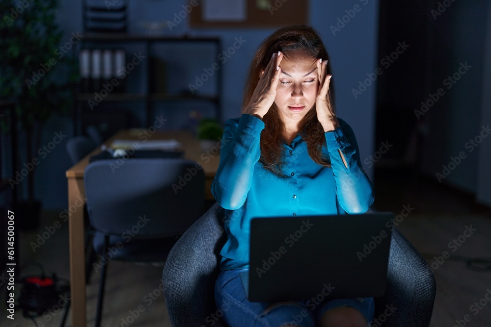 Brunette woman working at the office at night suffering from headache desperate and stressed because pain and migraine. hands on head. Brunette woman working at the office at night suffering from headache desperate and stressed because pain and migraine. hands on head.