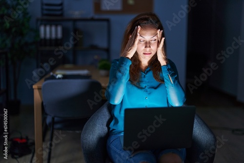Brunette woman working at the office at night suffering from headache desperate and stressed because pain and migraine. hands on head.