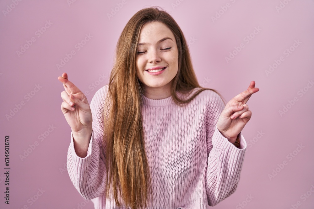 Young caucasian woman standing over pink background gesturing finger crossed smiling with hope and eyes closed. luck and superstitious concept. Young caucasian woman standing over pink background gesturing finger crossed smiling with hope and eyes closed. luck and superstitious concept.