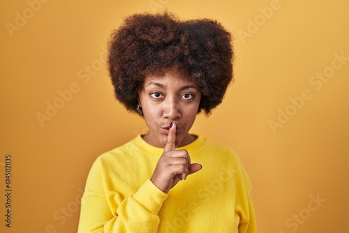 Young african american woman standing over yellow background asking to be quiet with finger on lips. silence and secret concept.