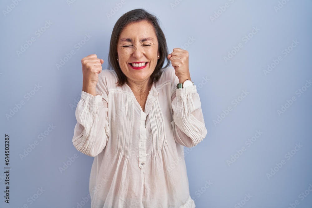 Middle age hispanic woman standing over blue background excited for success with arms raised and eyes closed celebrating victory smiling. winner concept. Middle age hispanic woman standing over blue background excited for success with arms raised and eyes closed celebrating victory smiling. winner concept.