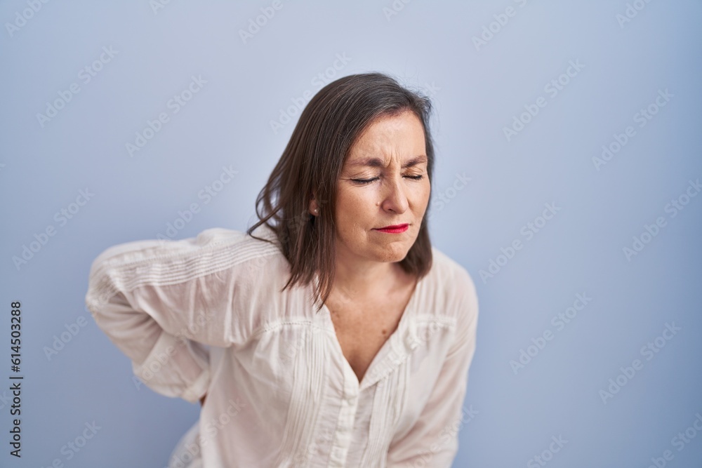 Middle age hispanic woman standing over blue background suffering of backache, touching back with hand, muscular pain Middle age hispanic woman standing over blue background suffering of backache, touching back with hand, muscular pain