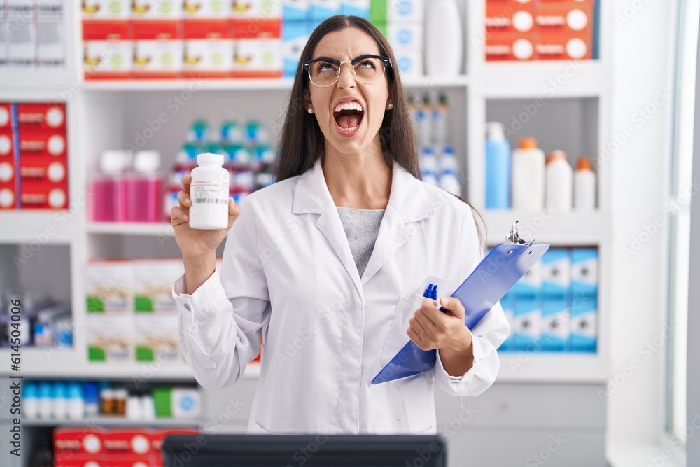 Young brunette woman working at pharmacy drugstore holding pills angry and mad screaming frustrated and furious, shouting with anger looking up.