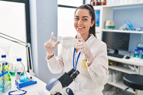 Young beautiful hispanic woman scientist smiling confident holding medical mask at laboratory