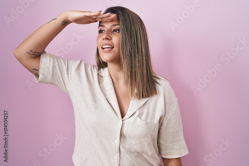 Blonde woman standing over pink background very happy and smiling looking far away with hand over head. searching concept.