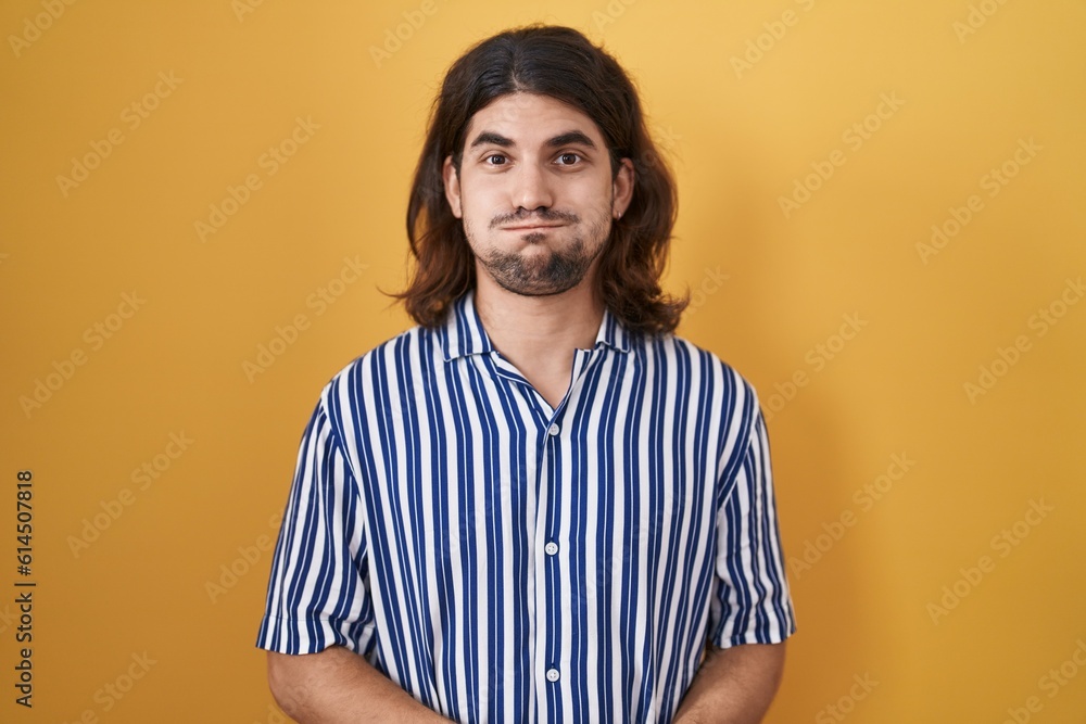 Hispanic man with long hair standing over yellow background puffing cheeks with funny face. mouth inflated with air, crazy expression.
