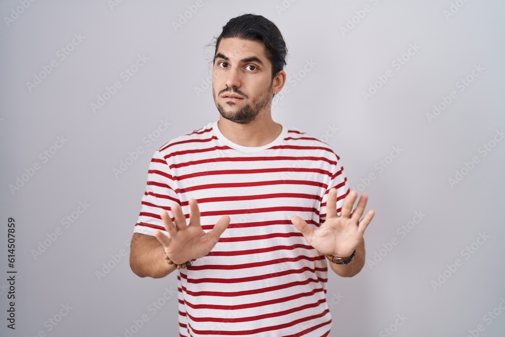 Hispanic man with long hair standing over isolated background moving away hands palms showing refusal and denial with afraid and disgusting expression. stop and forbidden. Hispanic man with long hair standing over isolated background moving away hands palms showing refusal and denial with afraid and disgusting expression. stop and forbidden.