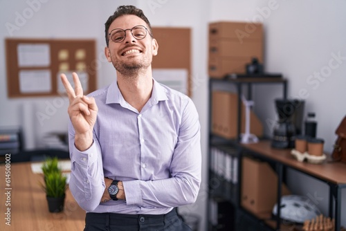 Young hispanic man at the office smiling with happy face winking at the camera doing victory sign with fingers. number two.