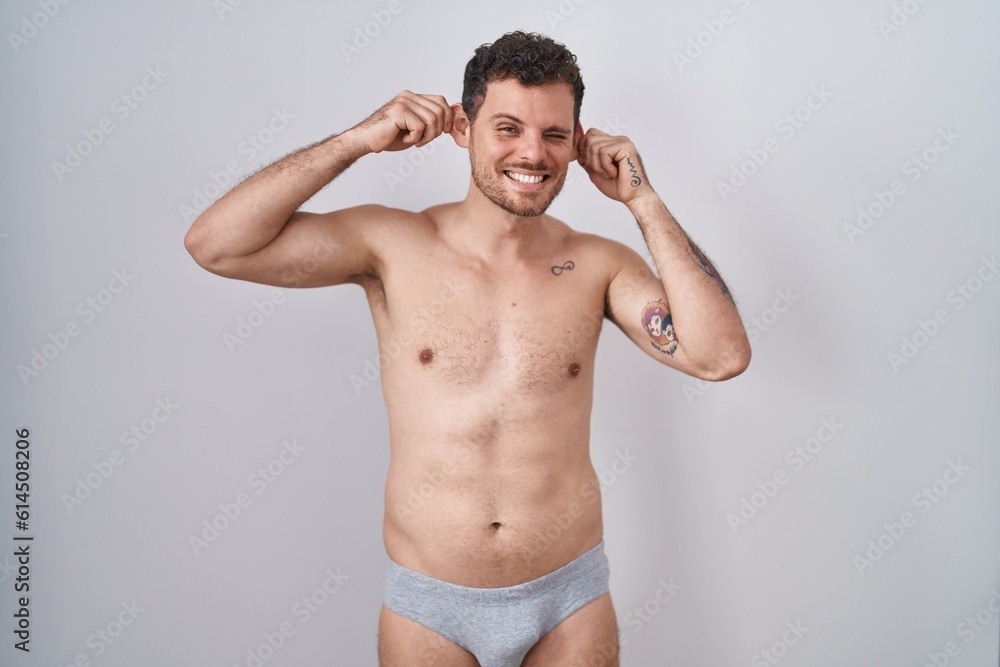 Young hispanic man standing shirtless wearing underware smiling pulling ears with fingers, funny gesture. audition problem
