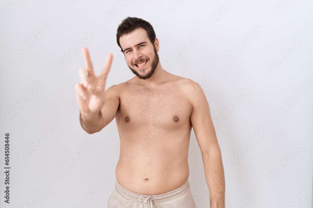 Young hispanic man standing shirtless over white background smiling looking to the camera showing fingers doing victory sign. number two.