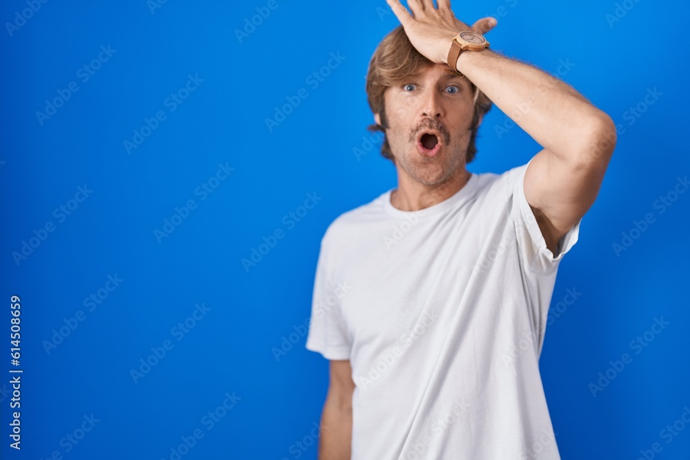 Naklejka premium Middle age man standing over blue background surprised with hand on head for mistake, remember error. forgot, bad memory concept.