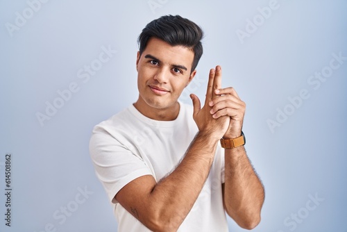 Hispanic man standing over blue background holding symbolic gun with hand gesture, playing killing shooting weapons, angry face