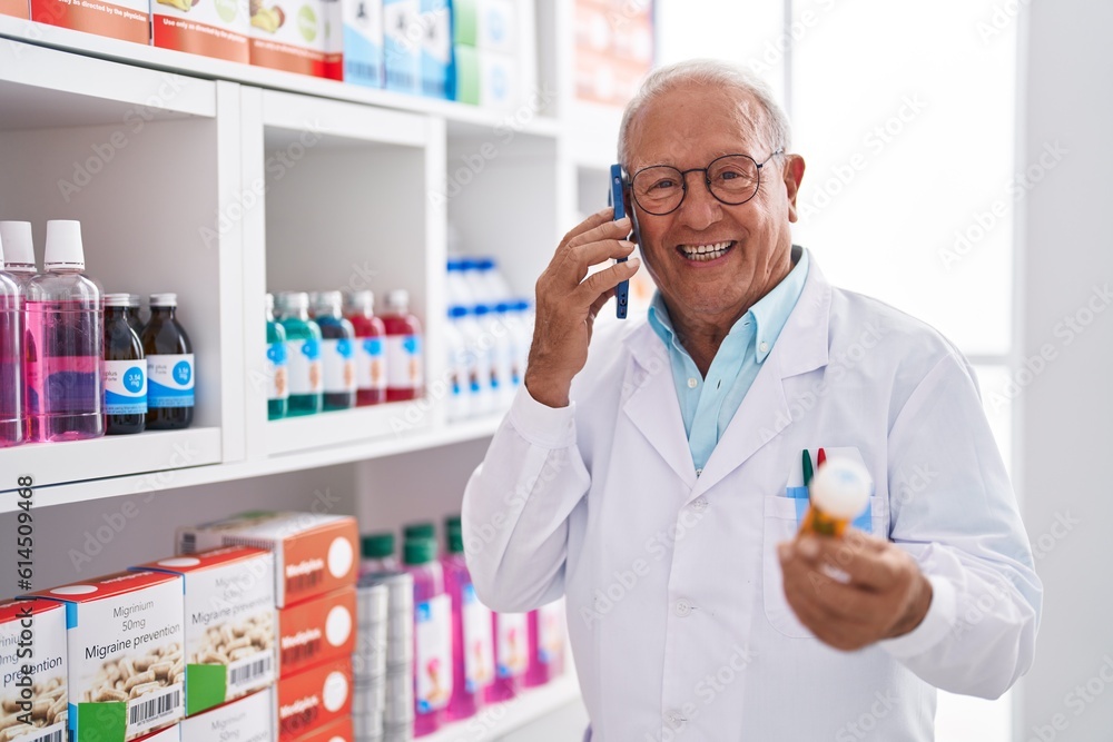 Senior grey-haired man pharmacist talking on smartphone holding pills at pharmacy