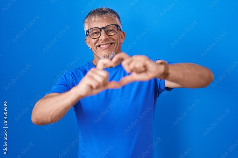 Hispanic man with grey hair standing over blue background smiling in love doing heart symbol shape with hands. romantic concept. Hispanic man with grey hair standing over blue background smiling in love doing heart symbol shape with hands. romantic concept.