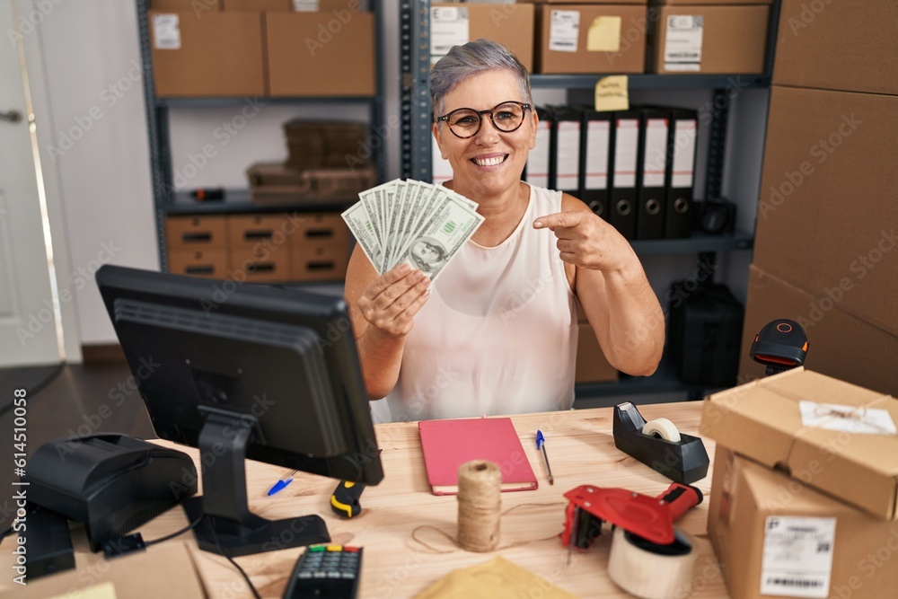 Middle age woman working at small business ecommerce holding dollars smiling happy pointing with hand and finger Middle age woman working at small business ecommerce holding dollars smiling happy pointing with hand and finger