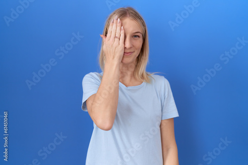 Young caucasian woman wearing casual blue t shirt covering one eye with hand, confident smile on face and surprise emotion.