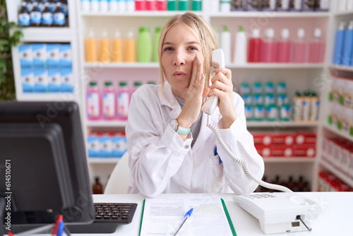 Young caucasian woman working at pharmacy drugstore speaking on the telephone hand on mouth telling secret rumor, whispering malicious talk conversation