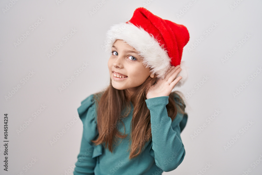 Little caucasian girl wearing christmas hat smiling with hand over ear listening and hearing to rumor or gossip. deafness concept. Little caucasian girl wearing christmas hat smiling with hand over ear listening and hearing to rumor or gossip. deafness concept.
