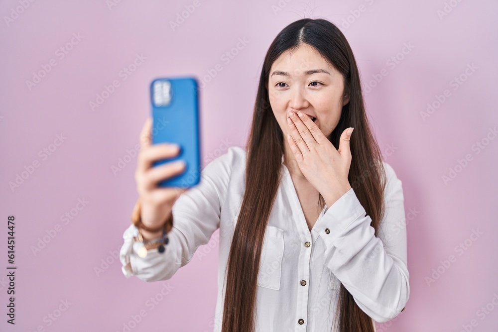Chinese young woman taking a selfie photo with smartphone covering mouth with hand, shocked and afraid for mistake. surprised expression Chinese young woman taking a selfie photo with smartphone covering mouth with hand, shocked and afraid for mistake. surprised expression