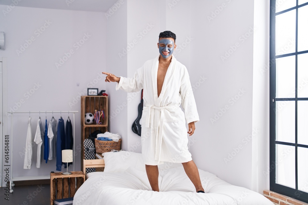 Young latin man dancing on bed with facial mask treatment at bedroom