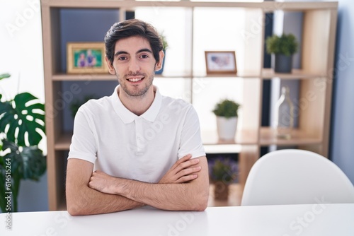Young hispanic man smiling confident sitting on table with arms crossed gesture at home