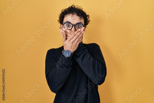 Hispanic man standing over yellow background shocked covering mouth with hands for mistake. secret concept.
