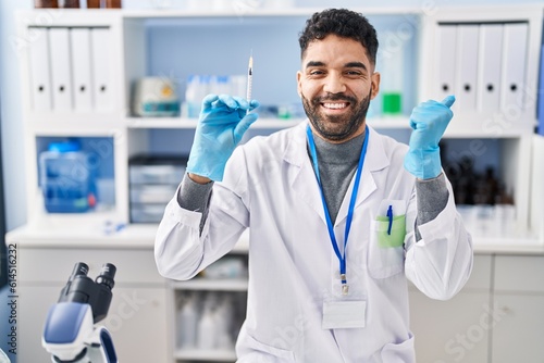 Hispanic man with beard working at scientist laboratory holding syringe screaming proud  celebrating victory and success very excited with raised arm