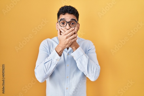 Hispanic man with beard standing over yellow background shocked covering mouth with hands for mistake. secret concept.