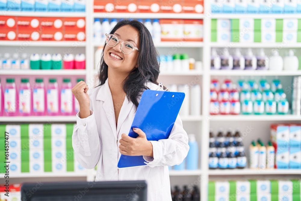 Asian young woman working at pharmacy drugstore holding clipboard screaming proud, celebrating victory and success very excited with raised arms