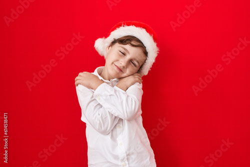 Little caucasian boy wearing christmas hat over red background hugging oneself happy and positive, smiling confident. self love and self care