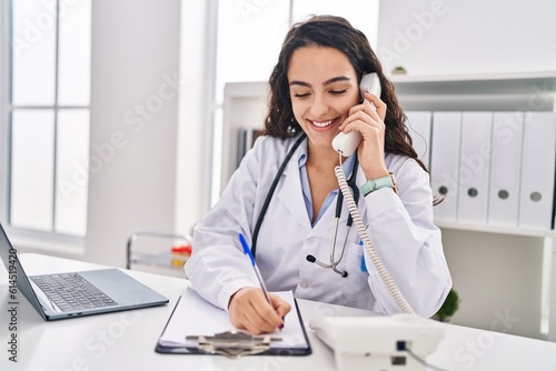 Young hispanic woman wearing doctor uniform talking on the telephone at clinic