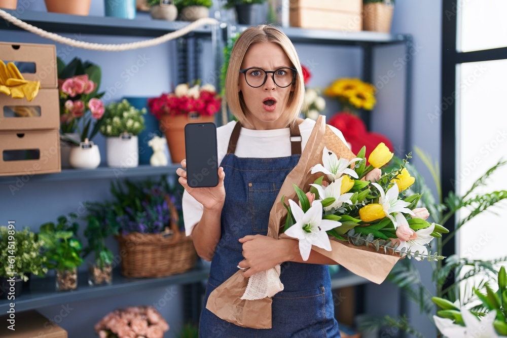 Young caucasian woman working at florist shop showing smartphone screen in shock face, looking skeptical and sarcastic, surprised with open mouth