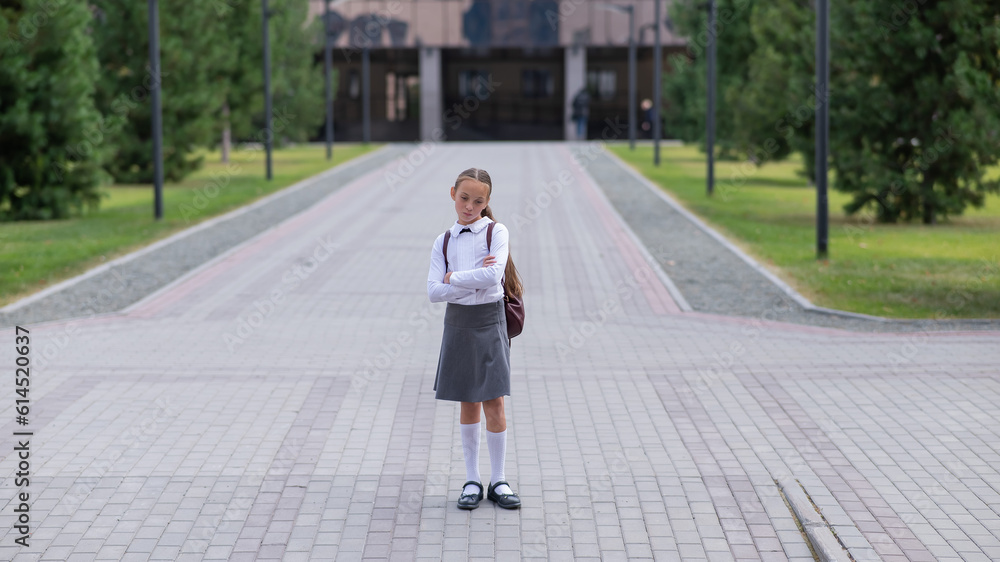 A sad caucasian girl in uniform and with a backpack holds her arms crossed on her chest stands outdoors after school. A sad caucasian girl in uniform and with a backpack holds her arms crossed on her chest stands outdoors after school.