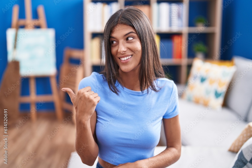 Brunette young woman sitting on the sofa at home smiling with happy face looking and pointing to the side with thumb up. Brunette young woman sitting on the sofa at home smiling with happy face looking and pointing to the side with thumb up.