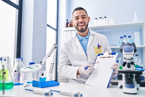 Hispanic man working at scientist laboratory holding blank clipboard smiling and laughing hard out loud because funny crazy joke.