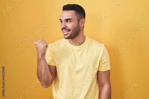 Young hispanic man standing over yellow background smiling with happy face looking and pointing to the side with thumb up. © Krakenimages.com