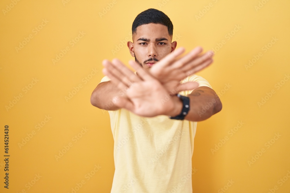 Young hispanic man standing over yellow background rejection expression crossing arms and palms doing negative sign, angry face