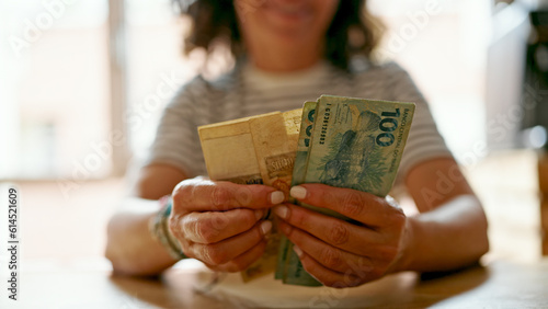 Middle age hispanic woman counting brazil real banknotes at home