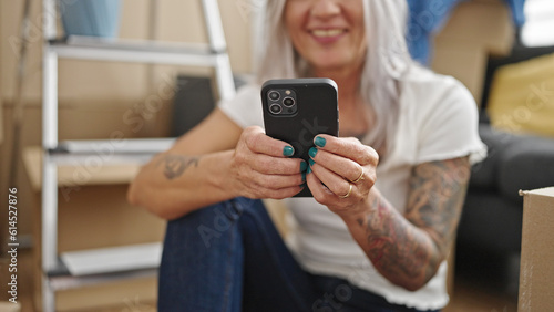Middle age grey-haired woman using smartphone sitting on floor at new home
